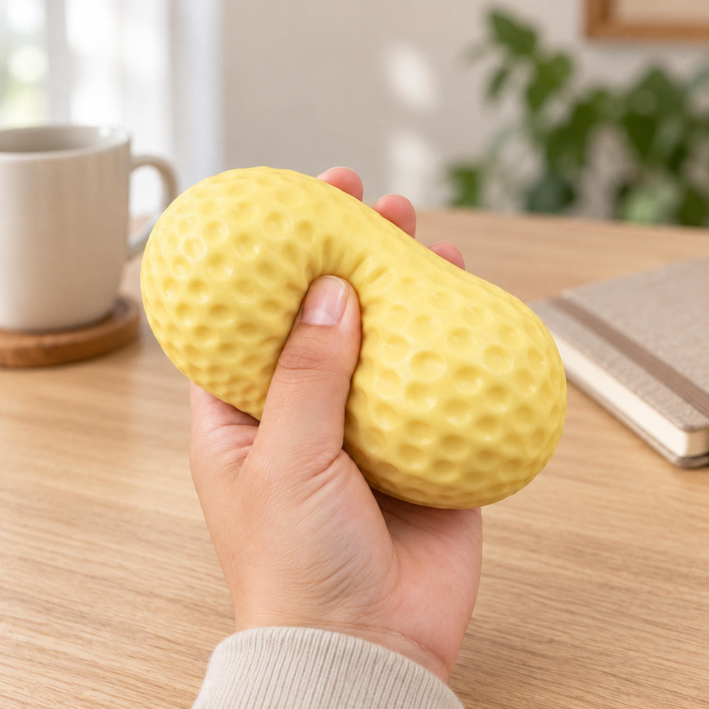 Hand squeezing a yellow peanut-shaped stress ball with a dimpled surface above a wooden desk.